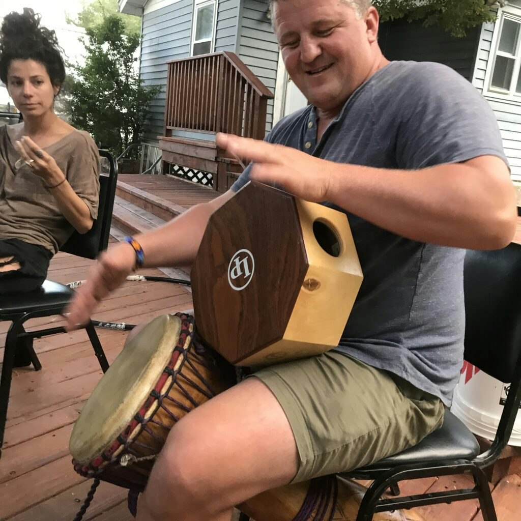 Two people sitting outdoors; one playing a drum, the other observing.