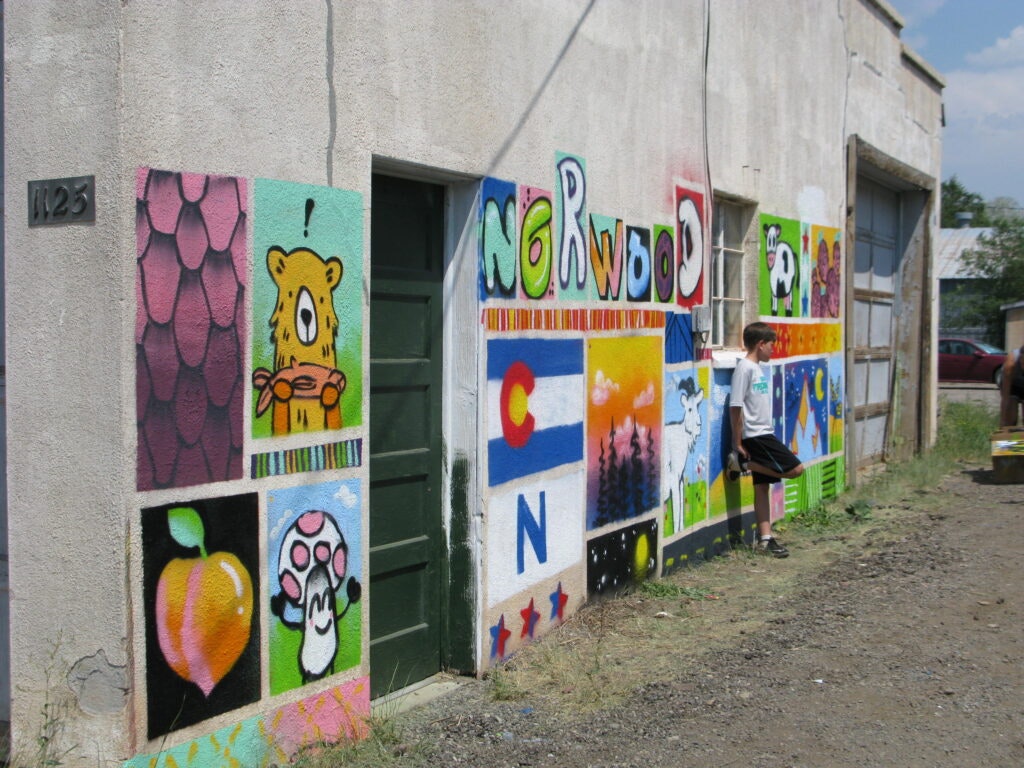A colorful mural on a building with various images, including a bear, mushrooms, and more, with a person standing nearby.