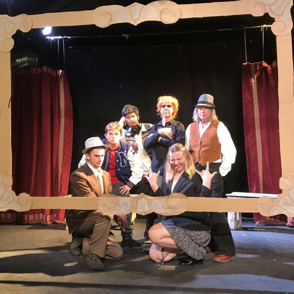 A group of people posing on stage within a decorative cardboard frame, with curtains in the background.