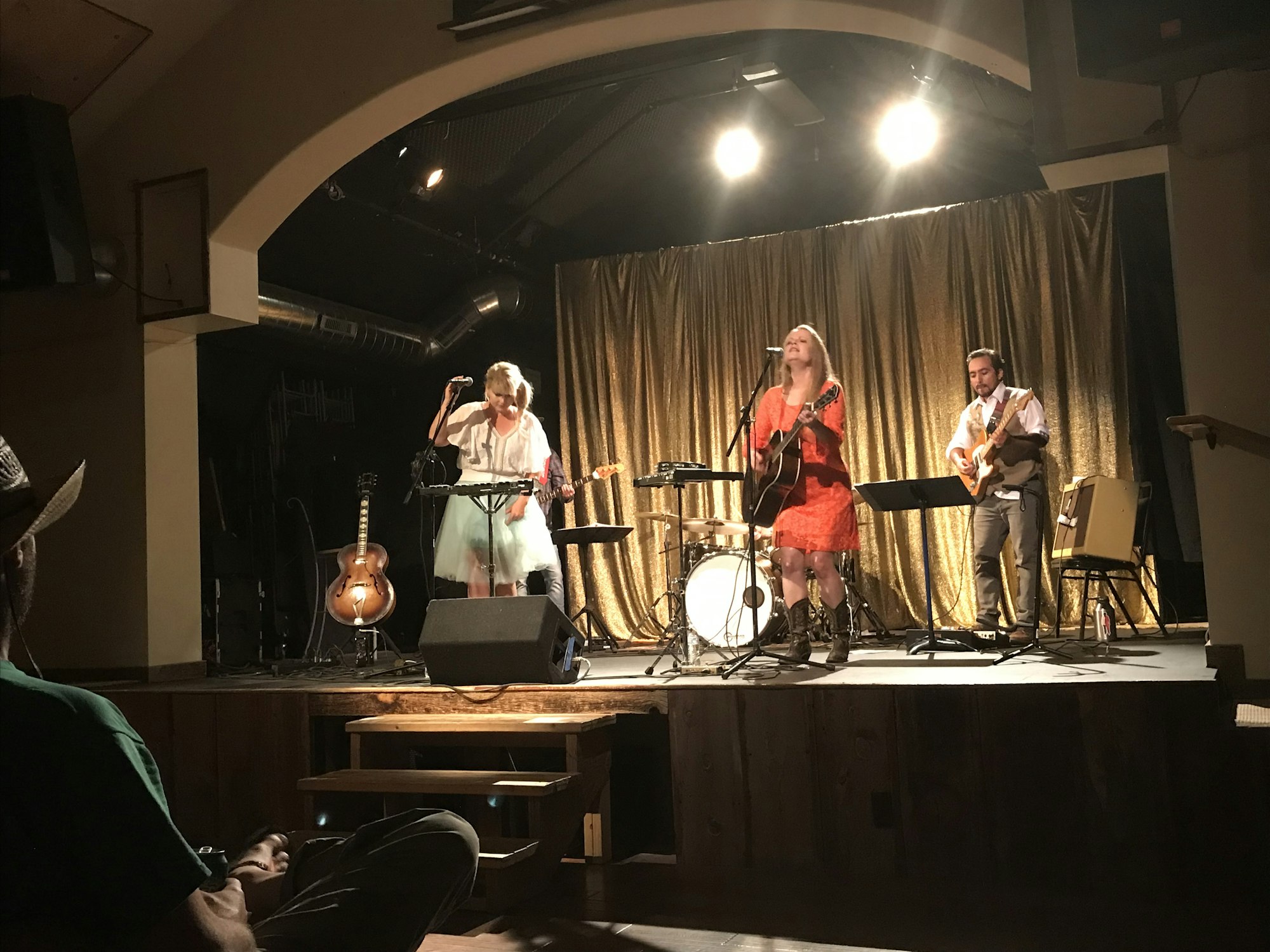 Three musicians perform on stage, playing guitars and a keyboard, with golden curtains as a backdrop.