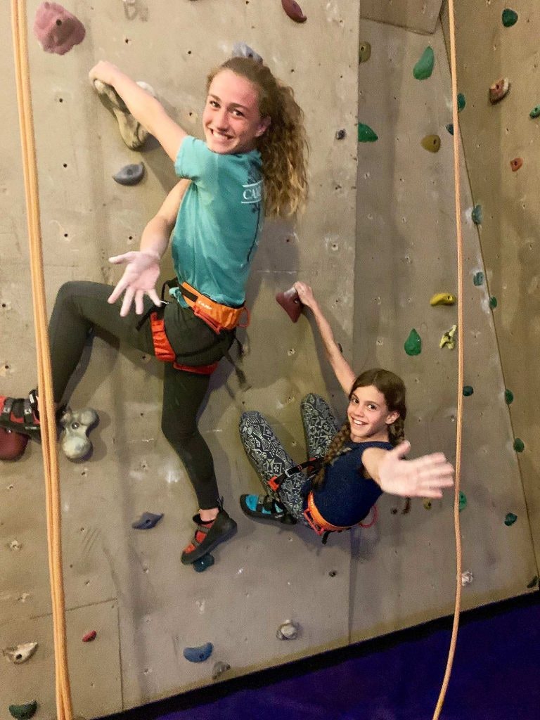 Two people climbing an indoor rock wall, smiling at the camera.