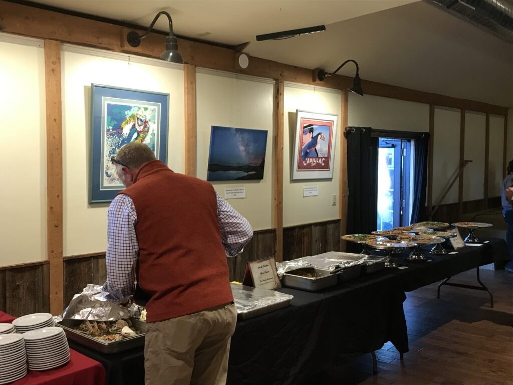 Buffet setup with food trays. A person serves themselves near framed artwork on the wall. Plates are stacked on the table.