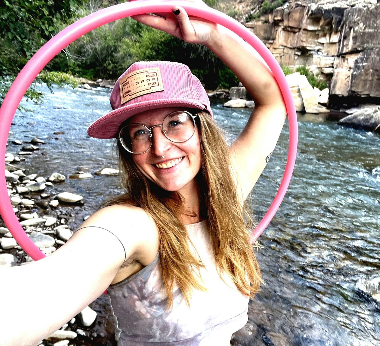 A person smiling by a river, holding a pink hula hoop and wearing glasses and a hat.