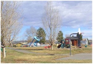 A playground with a slide, bare trees, grassy area, and a walking path under a cloudy sky.