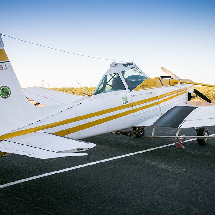 The image shows a small agricultural airplane parked on a tarmac, featuring yellow stripes and a propeller.
