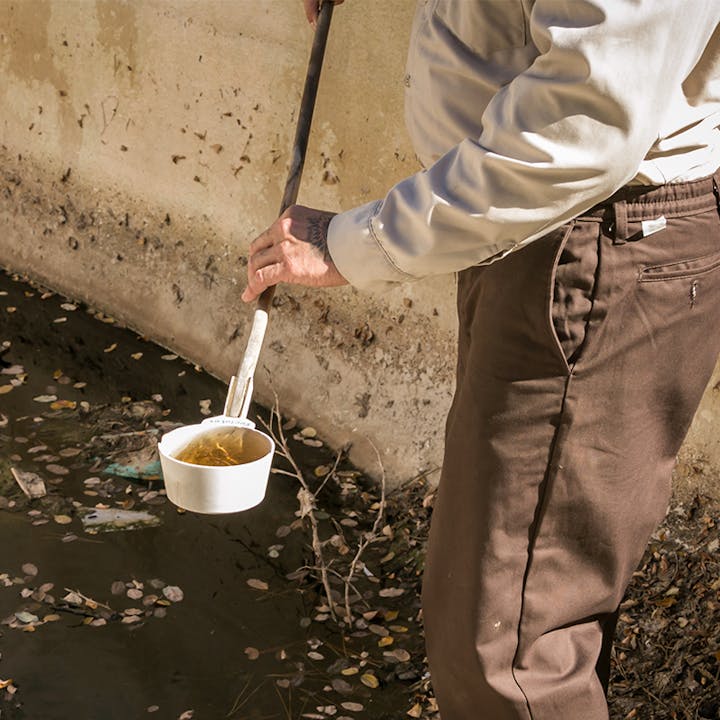 An individual is holding a container with liquid, possibly sampling water from a muddy area, with debris around.