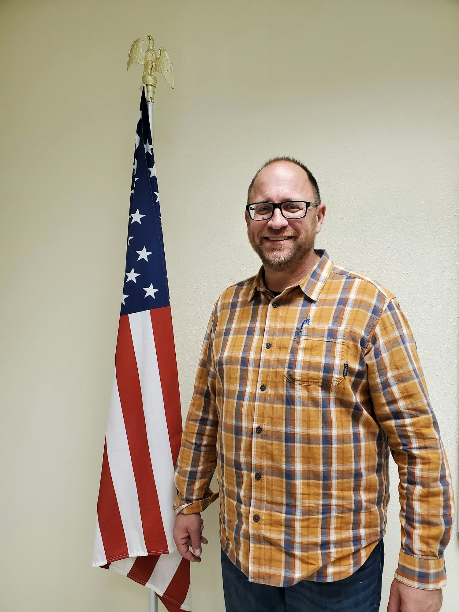 A person in a plaid shirt stands next to an American flag with a gold eagle topper.