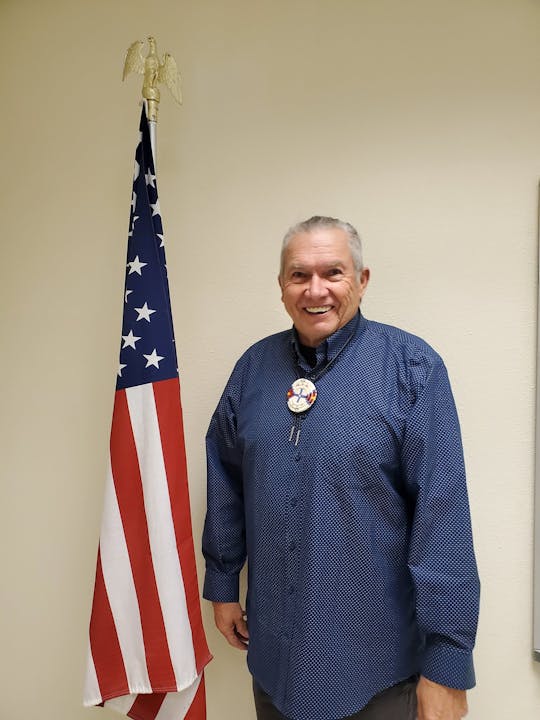 A person smiling next to an American flag inside a room, wearing a blue shirt with a bolo tie.