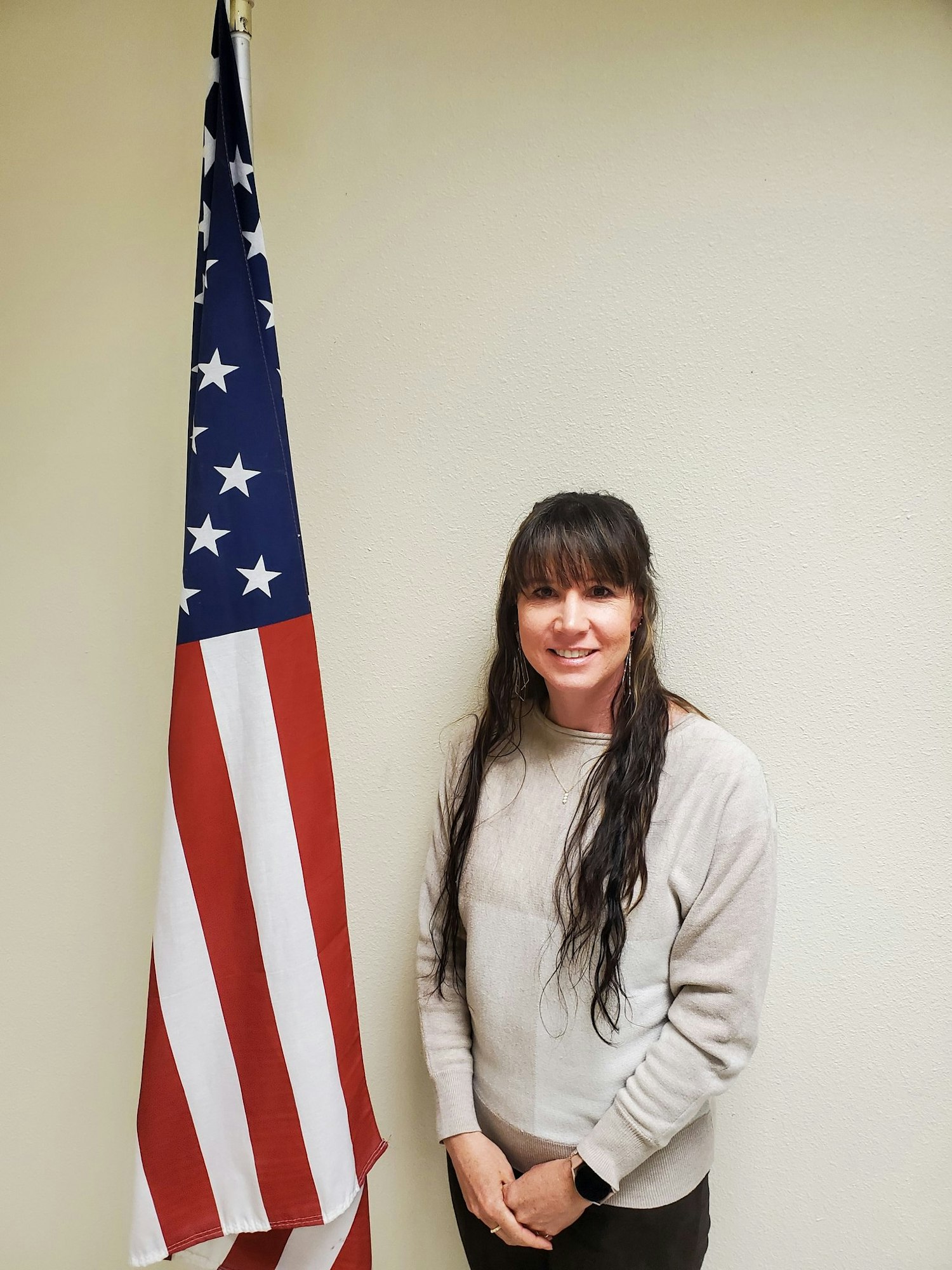 A woman standing next to an American flag, wearing a light sweater.