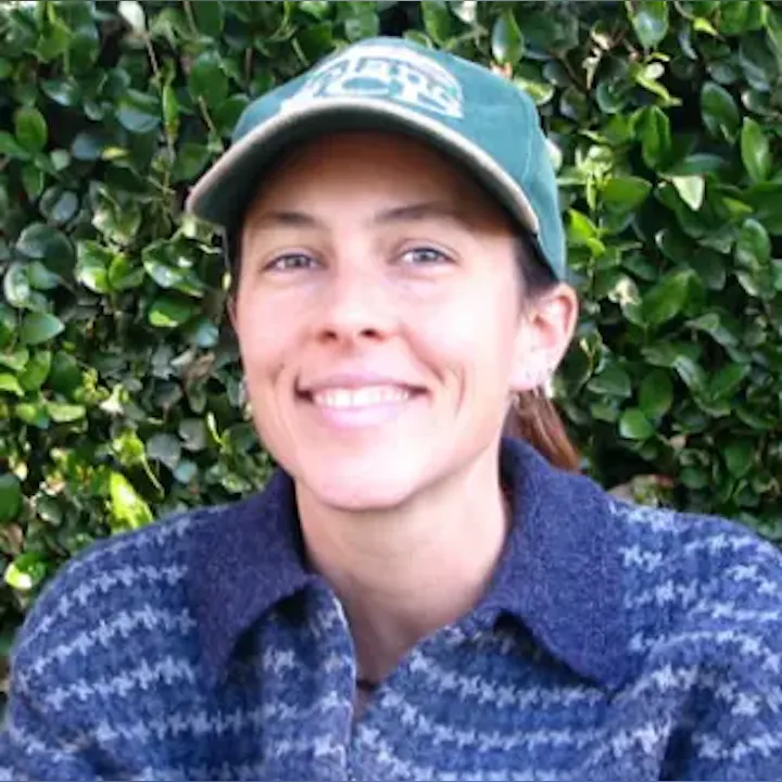A smiling person wearing a cap and a patterned jacket, with green foliage in the background.