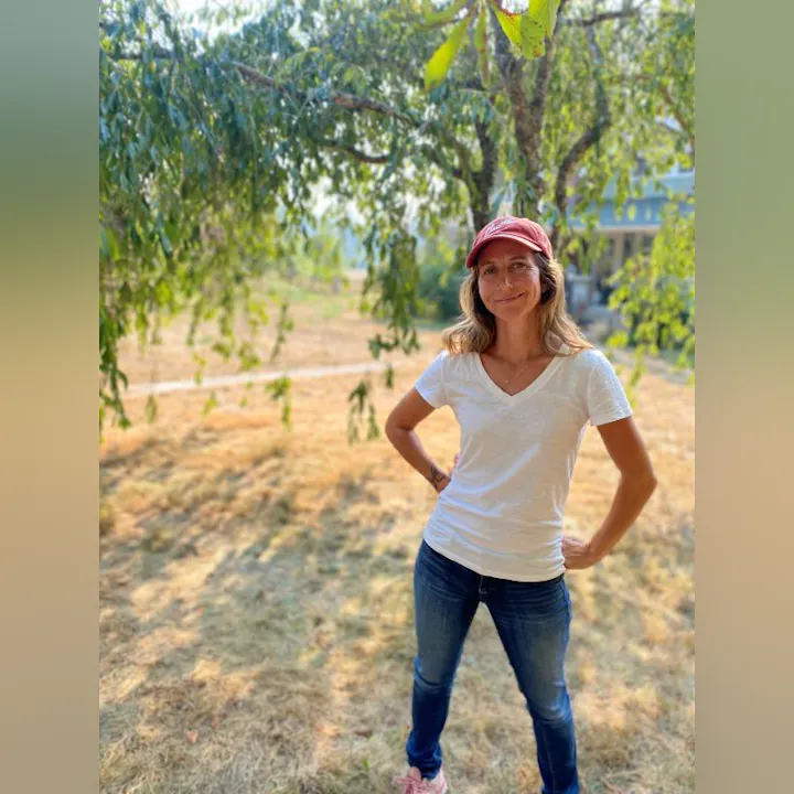 A smiling person stands outdoors under a tree, wearing a red cap and a white t-shirt, with a dry, grassy background.