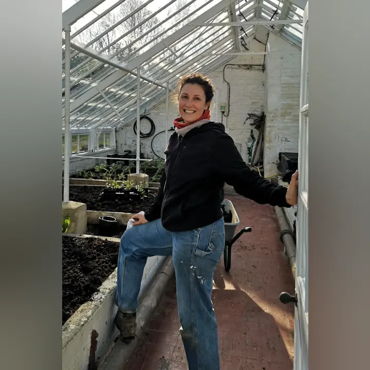 A smiling person stands in a greenhouse, wearing casual clothes and a scarf, with plants and gardening tools around.