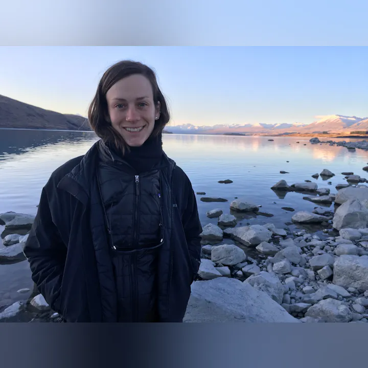 A smiling person stands by a serene lake surrounded by rocks, with mountains in the background during a clear day.