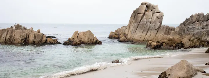 A serene coastal scene with rocky formations, gentle waves, and a calm sea under a gray sky.