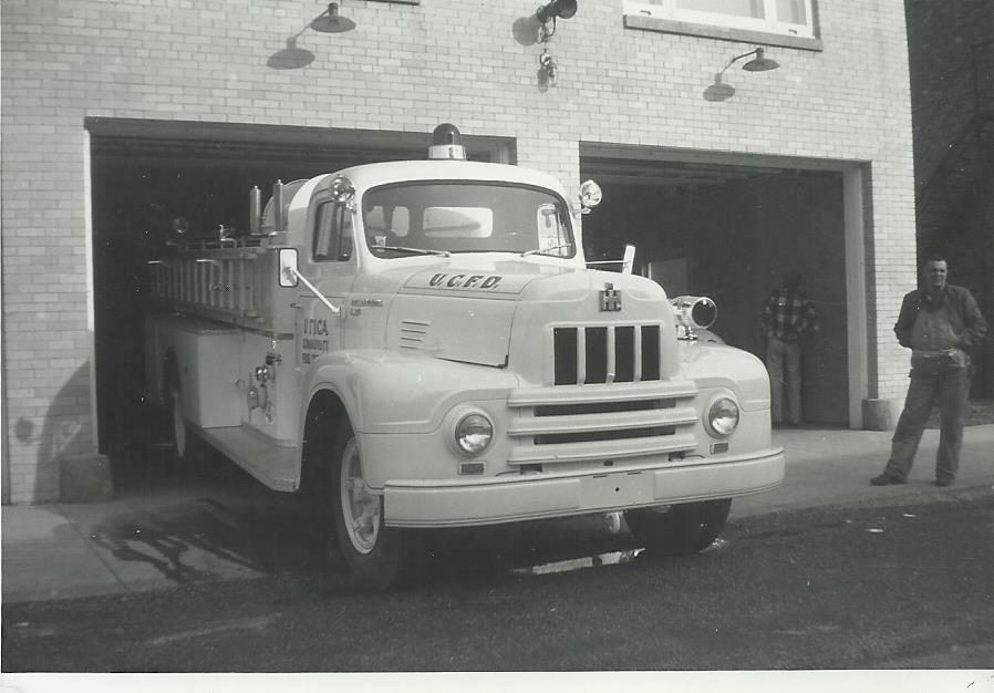A vintage white fire truck parked in front of a fire station, with a person standing nearby. The scene is in black and white.