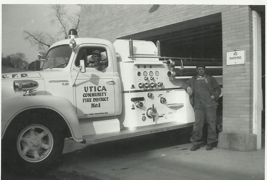 A vintage photo of firemen beside a fire truck labeled "Utica Community Fire District No. 1," with a "No Parking" sign nearby.