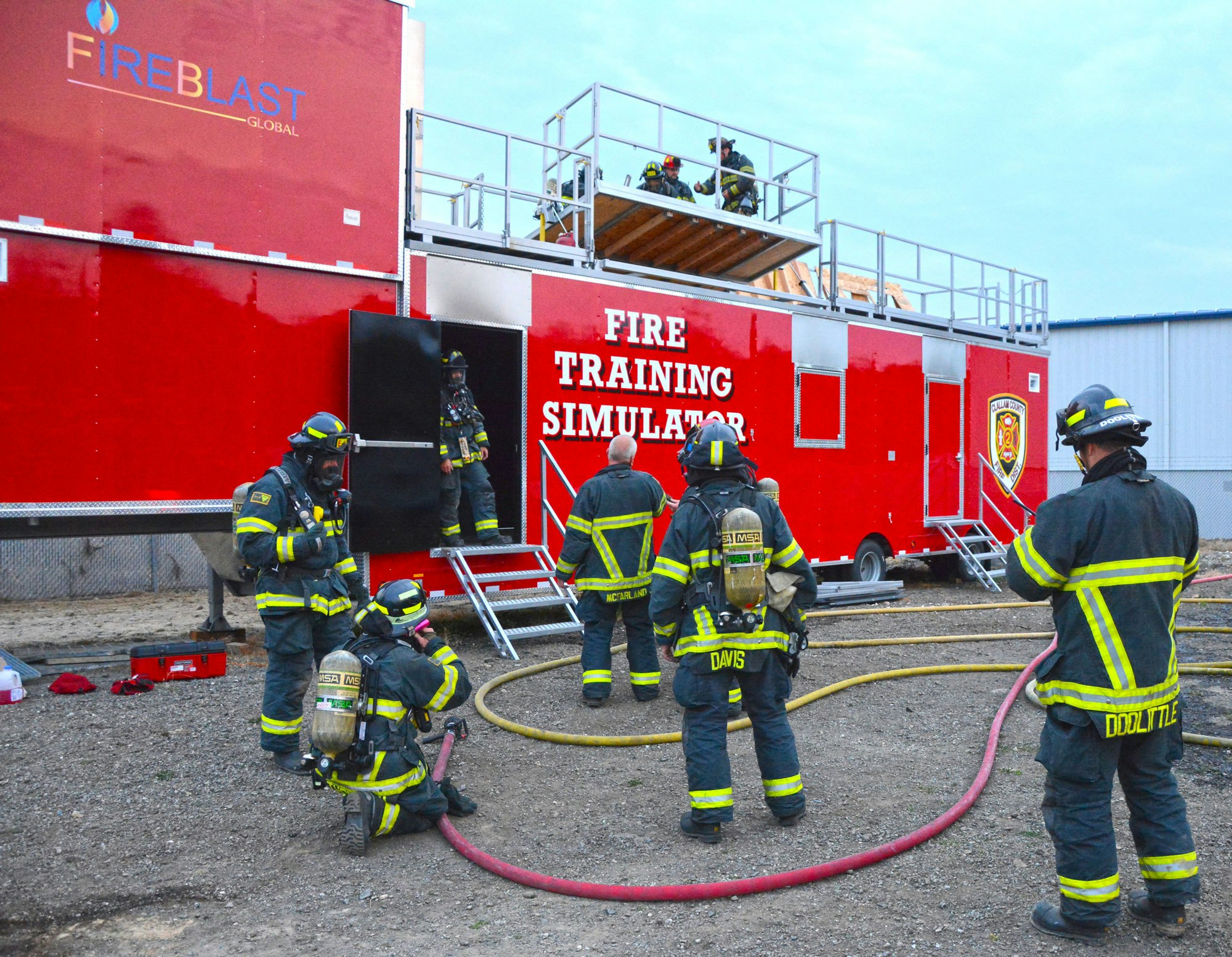 Firefighters training near a "Fire Training Simulator" with hoses and gear in a red truck setup.