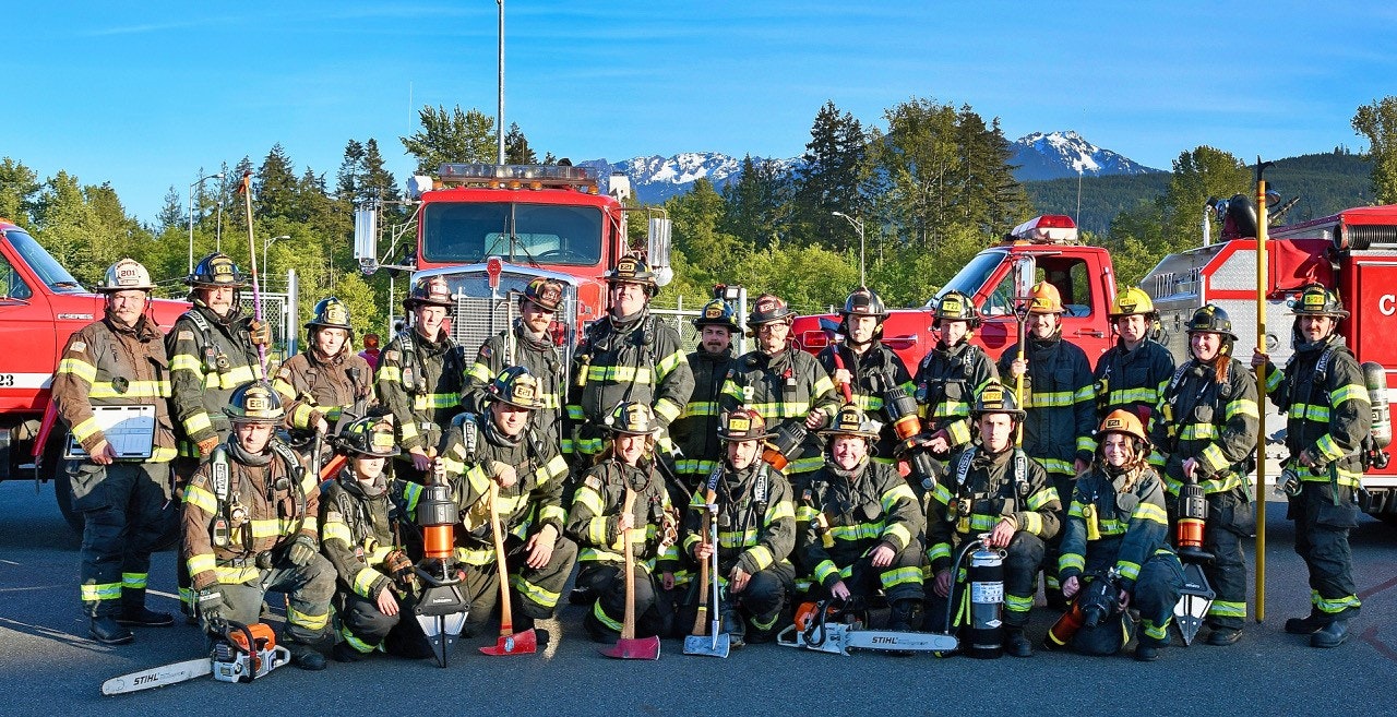 A group of firefighters in full gear in front of fire trucks with a mountain backdrop.