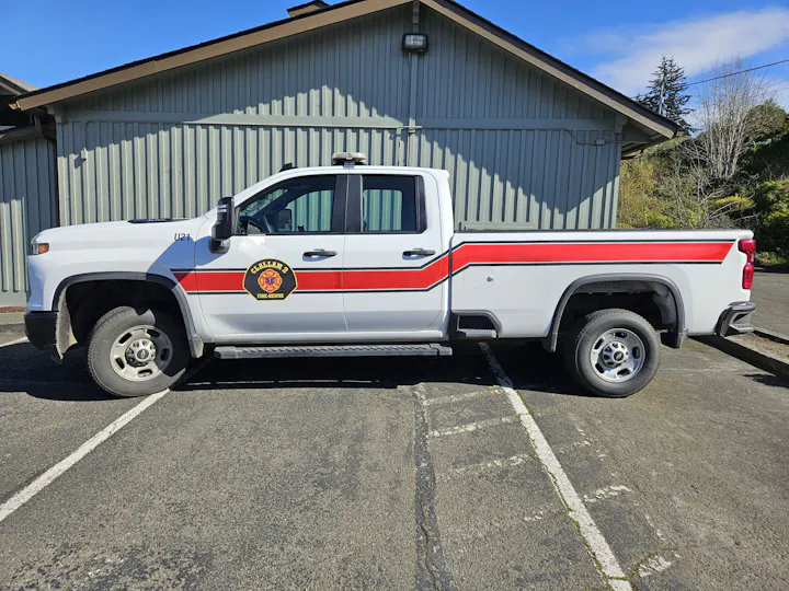 The image shows a white fire department pickup truck with a red stripe and a badge parked outside a building.