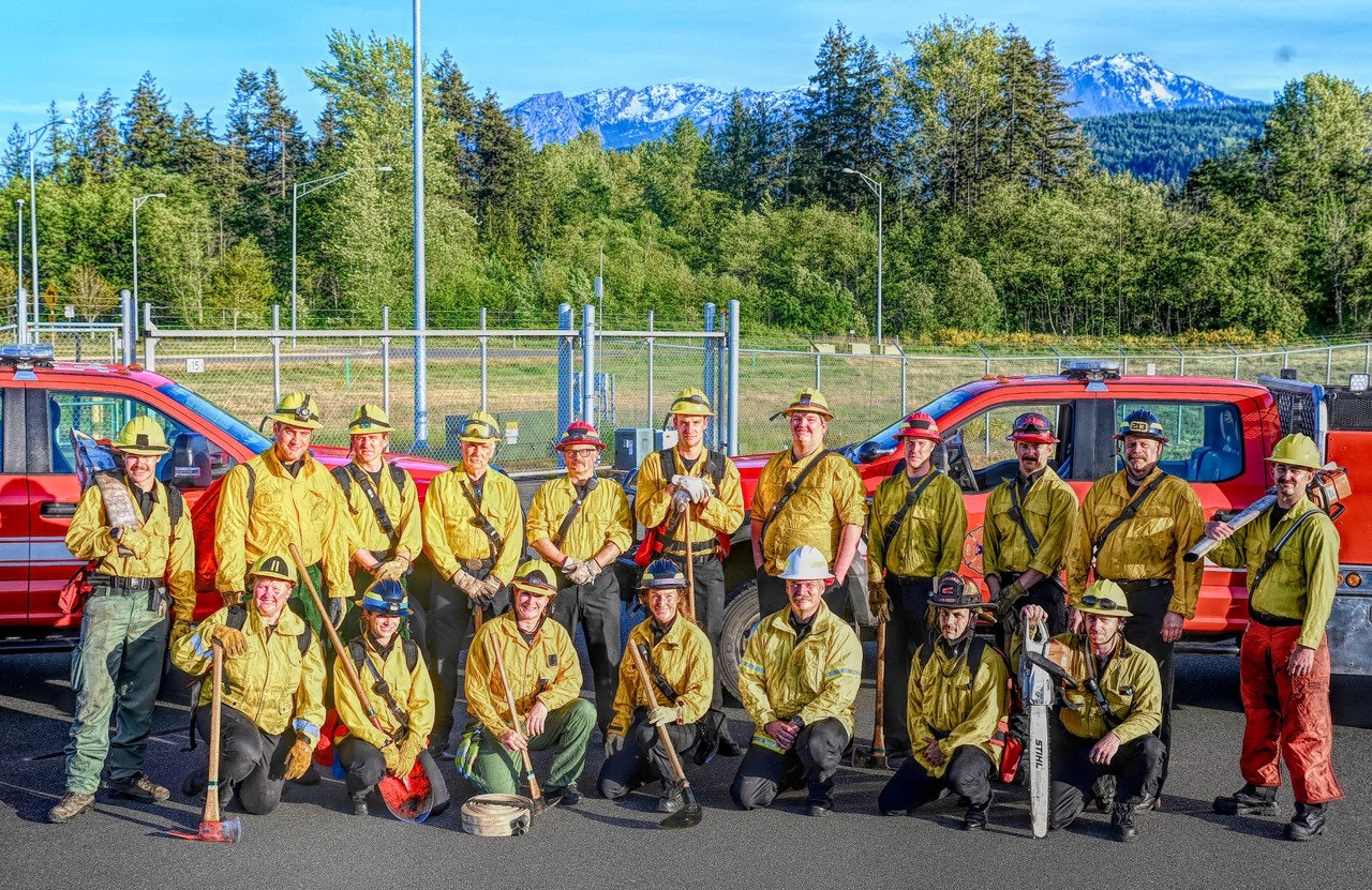 Group of firefighters in yellow shirts and helmets, with a fire truck and mountains in the background.