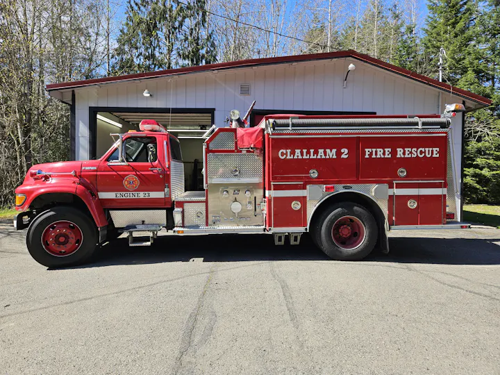 A red fire truck labeled "CLALLAM 2 FIRE RESCUE" parked in front of a building, surrounded by trees on a sunny day.