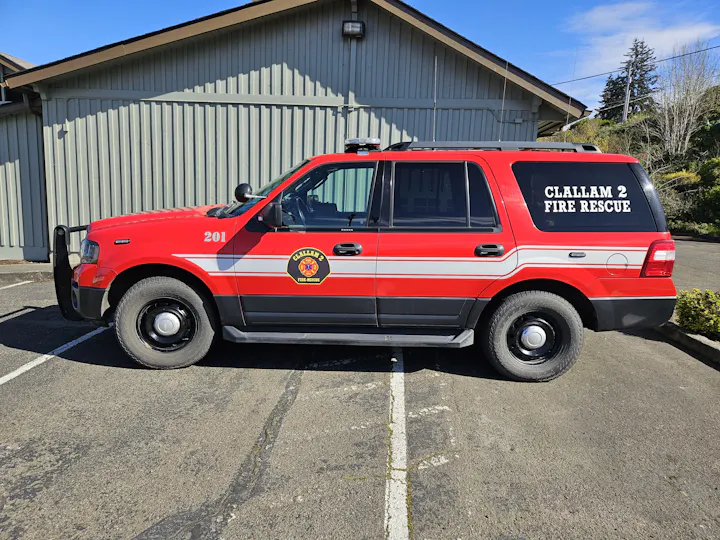 The image shows a red fire rescue vehicle labeled "CLALLAM 2" parked beside a building.