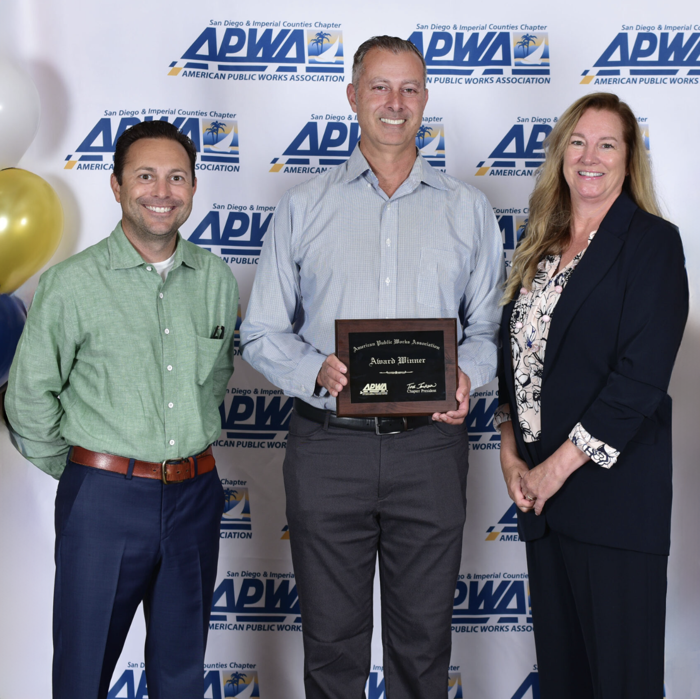 Three people standing in front of an APWA backdrop; the middle person holds an award plaque.