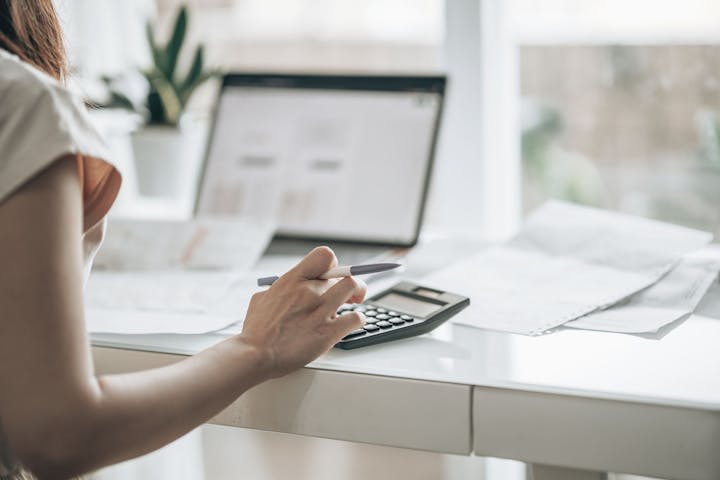 A person is using a calculator and holding a pen while reviewing documents at a desk with a laptop and a plant in the background.