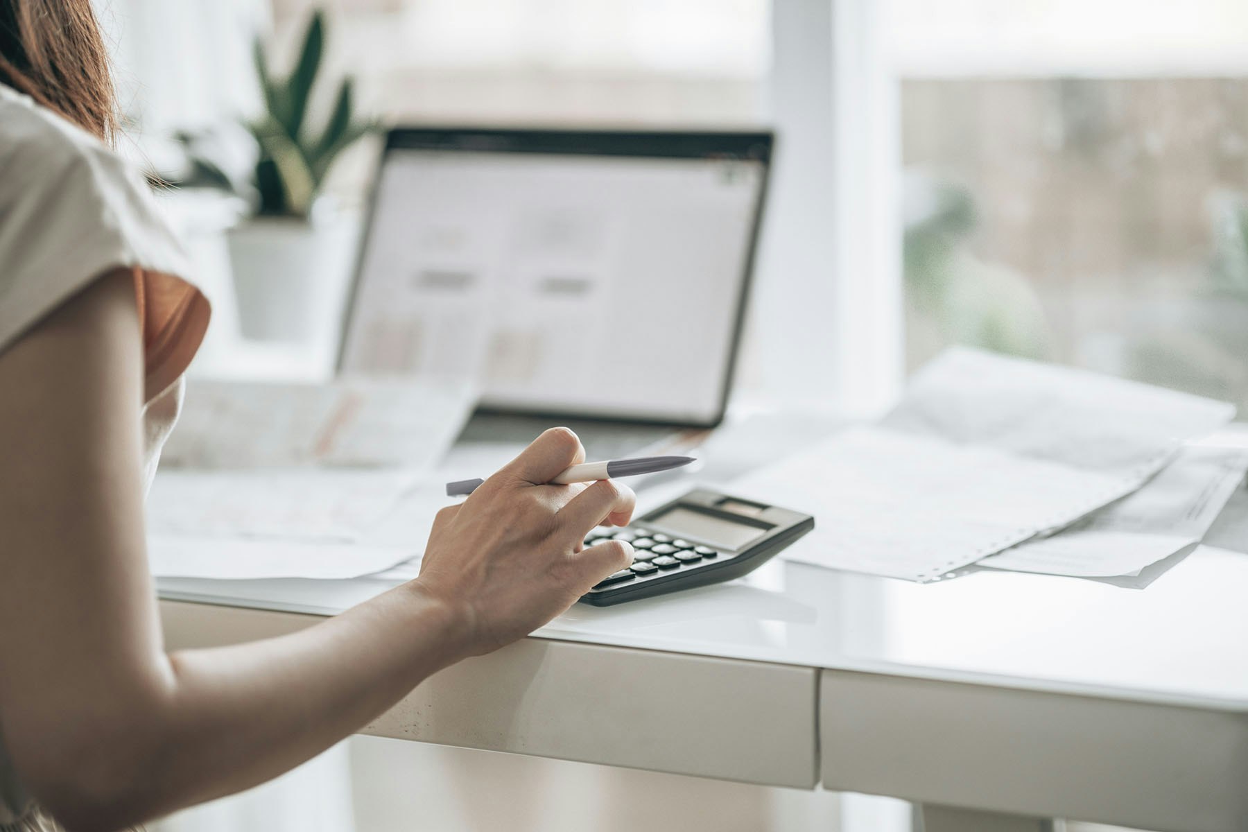 A person is using a calculator and holding a pen while reviewing documents at a desk with a laptop and a plant in the background.