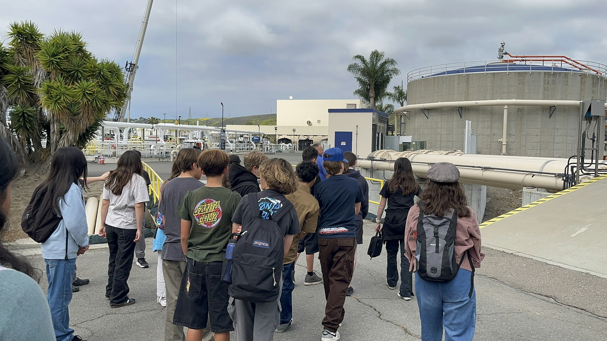 A group of people walking near industrial pipes and tanks outdoors.