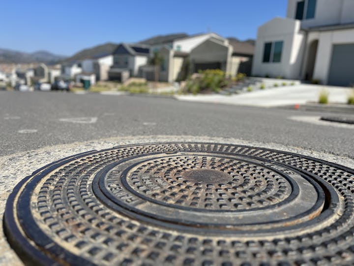 Close-up of a manhole cover on a street with blurred suburban houses in the background.