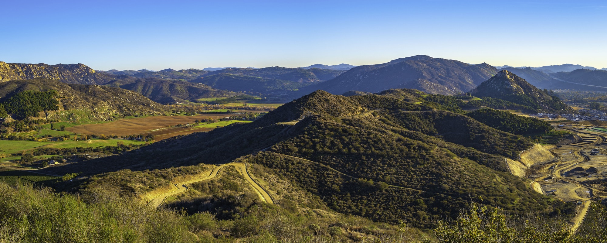 A panoramic landscape of Monserate Mountain Preserve showing rolling hills, winding roads, cultivated fields, and a clear blue sky.