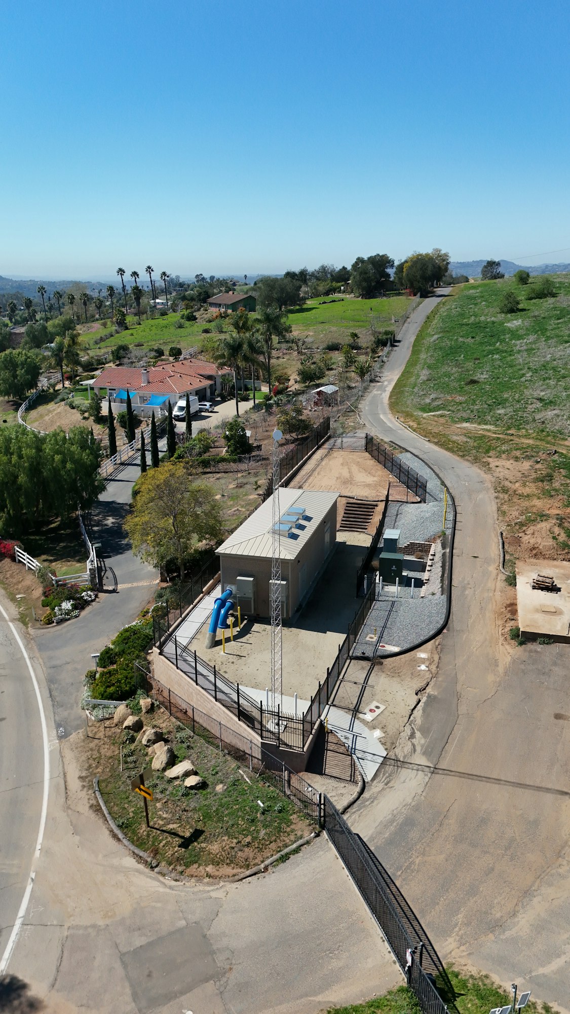 Aerial view of a utility building and surrounding land, fenced area, winding pathways, and nearby residential properties.