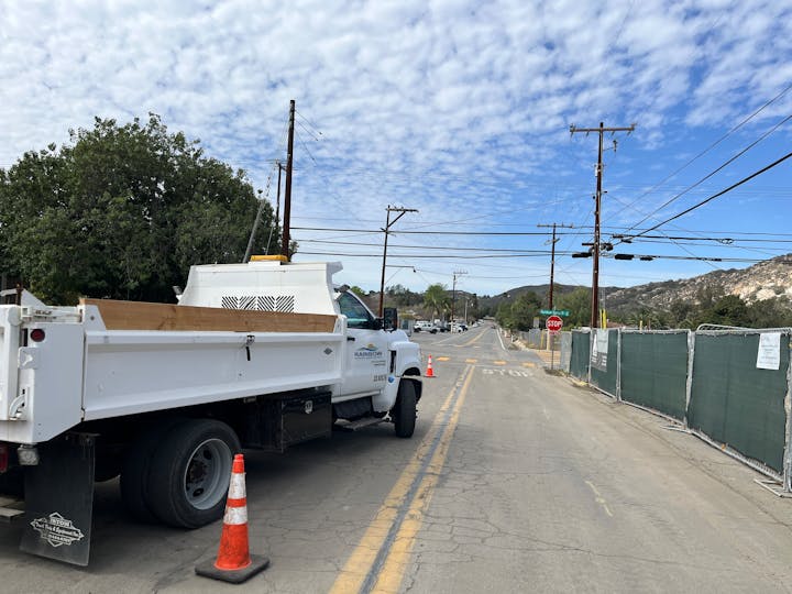 A white truck blocking a road with cones, surrounded by power lines, trees, and a construction fence under a cloudy sky.