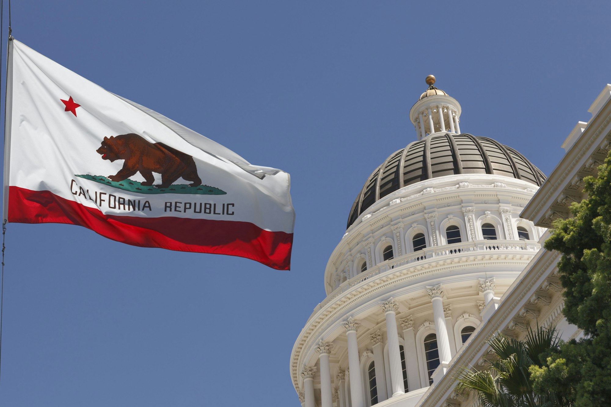 A California state flag waves in front of a grand dome building against a clear blue sky.