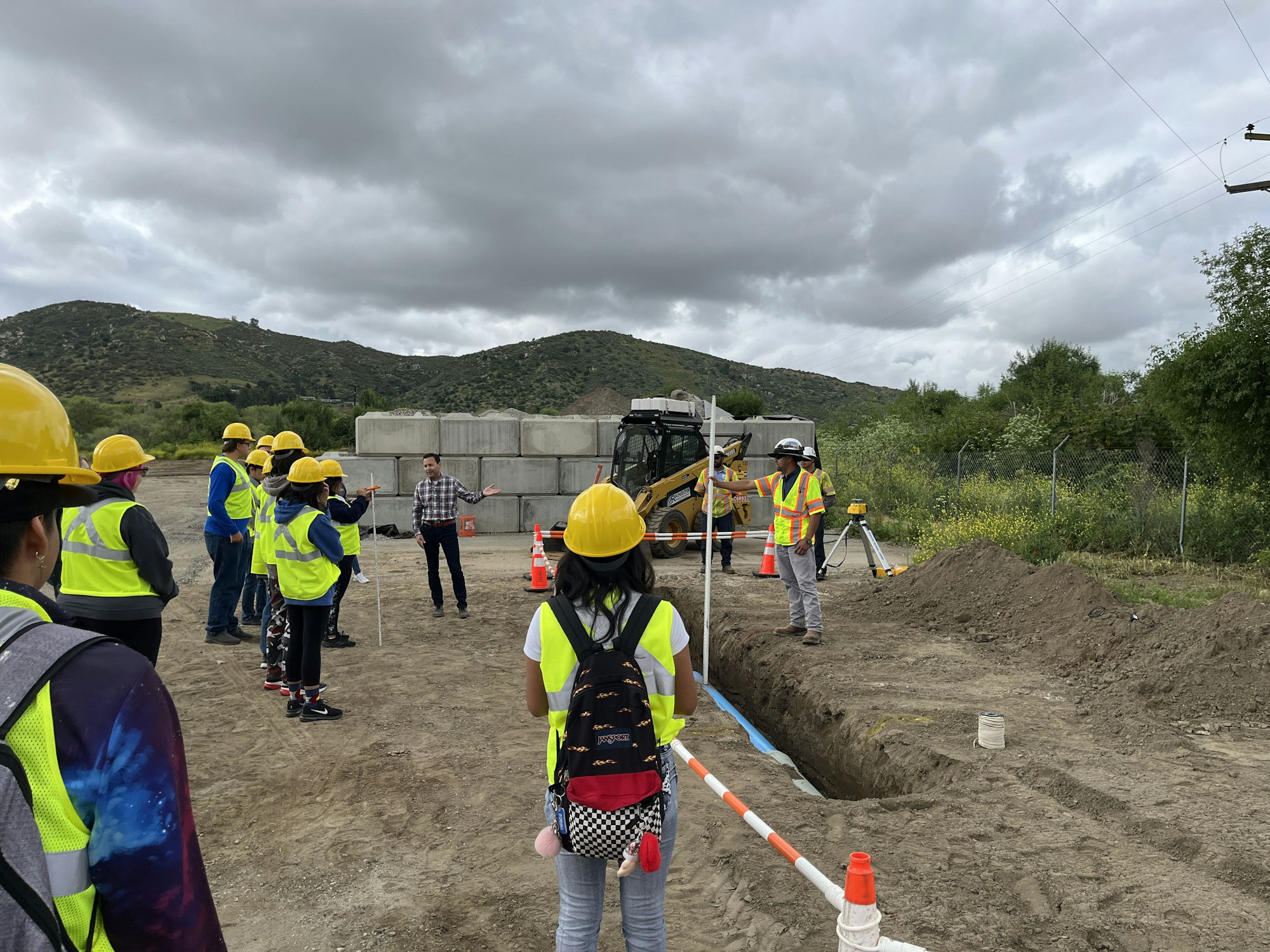 Group of people in hard hats at a construction site with an excavator and a trench in the background.