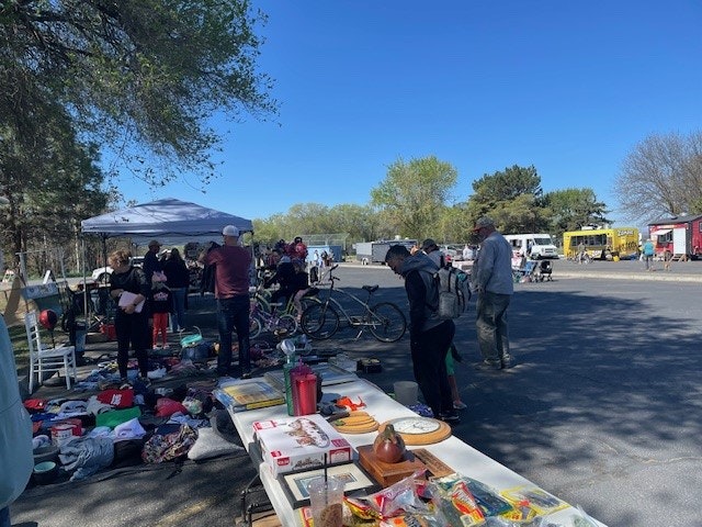 A bustling outdoor market scene with people browsing items, food vendors, and a sunny sky. Items include clothing and kitchenware.