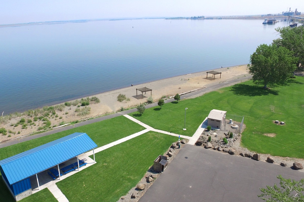 Aerial view of a lakeside park with a pavilion, walking paths, grass, and industrial ports in the distance.