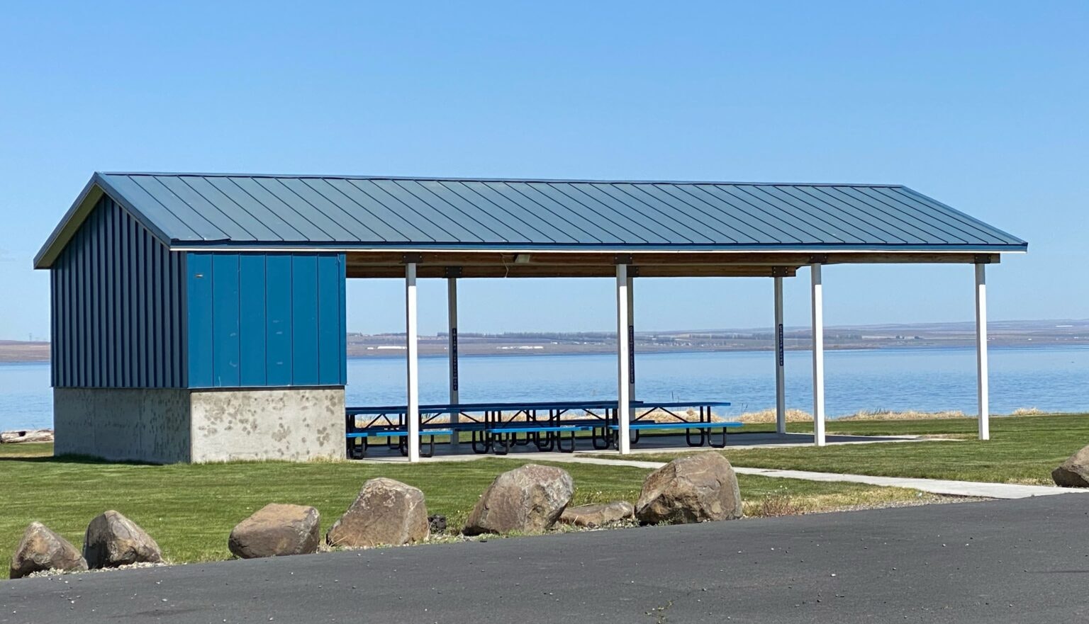A pavilion with picnic tables by a lake, clear skies, and rocks in the foreground.
