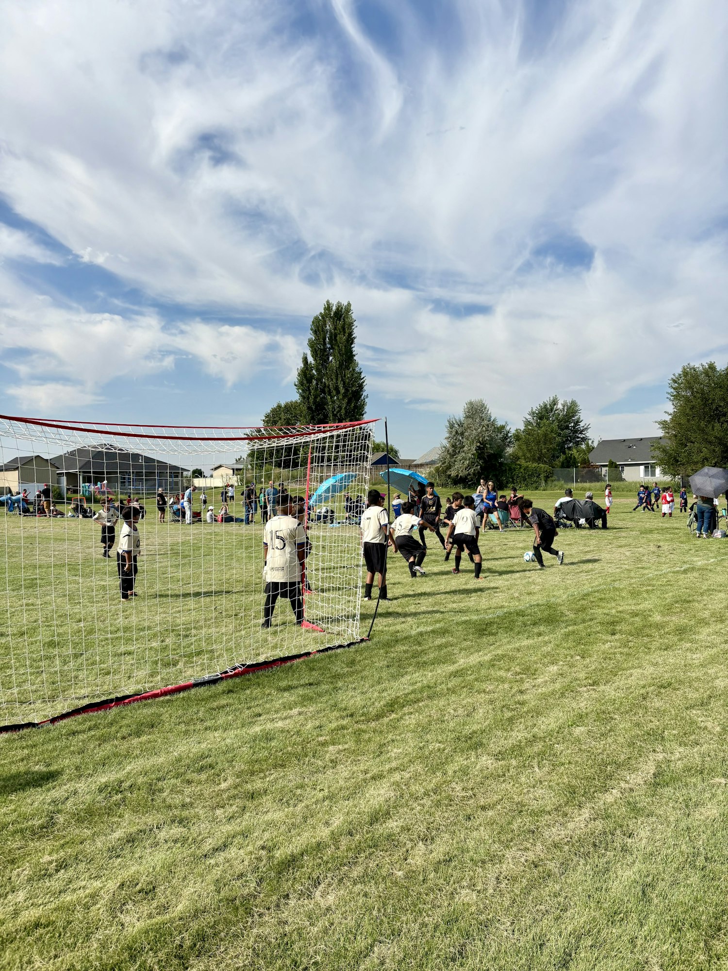Kids playing soccer on a grassy field, with a soccer net and spectators in the background under a partly cloudy sky.