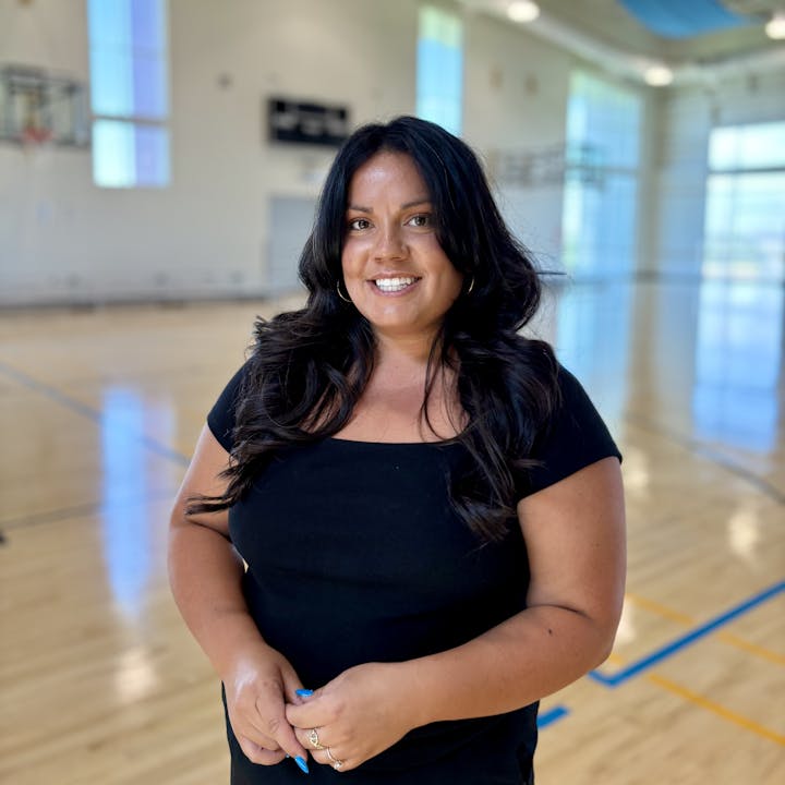 A person with long hair smiling, standing in a gymnasium with a basketball court.