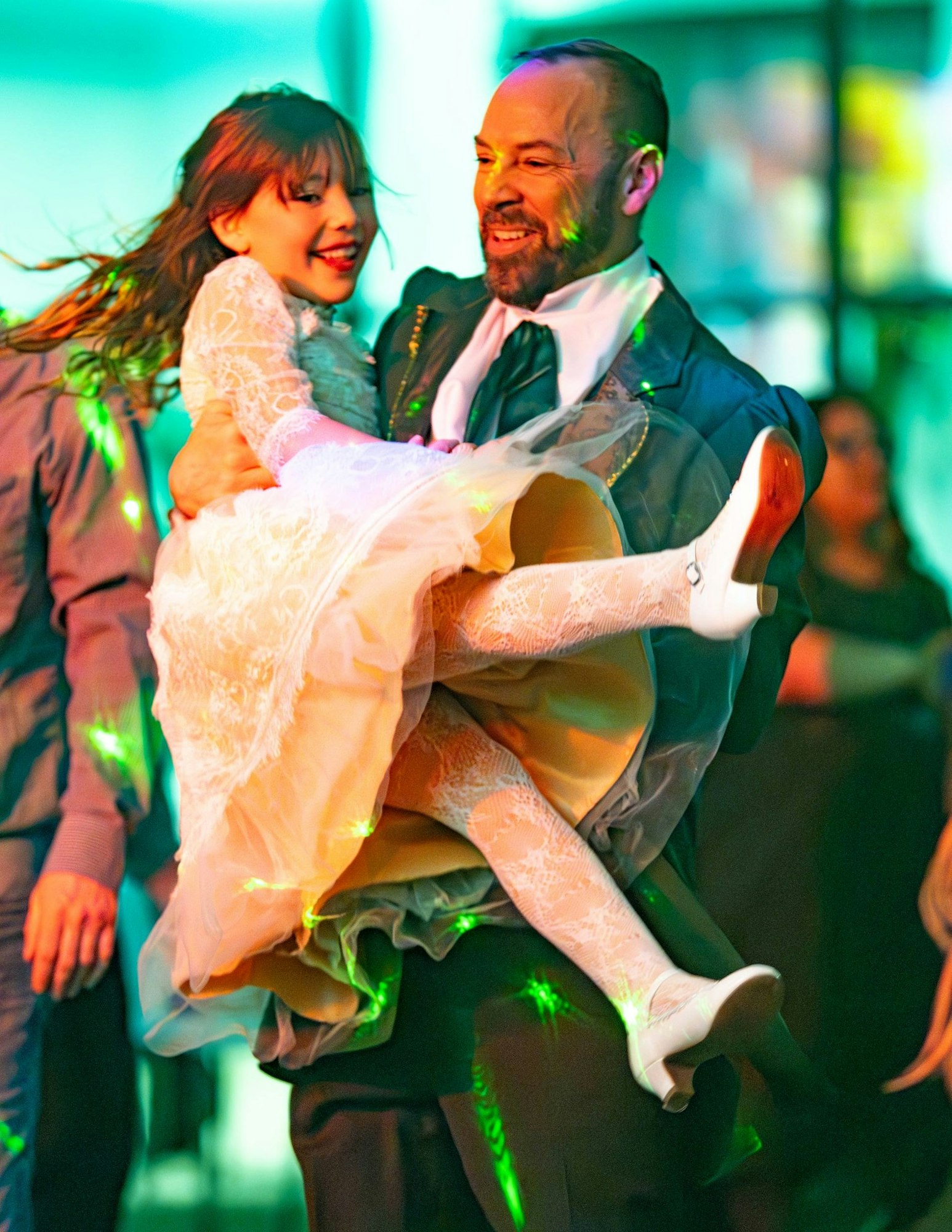 A man is happily lifting a girl in a dress, surrounded by colorful lights, as they enjoy a dance at a festive event.