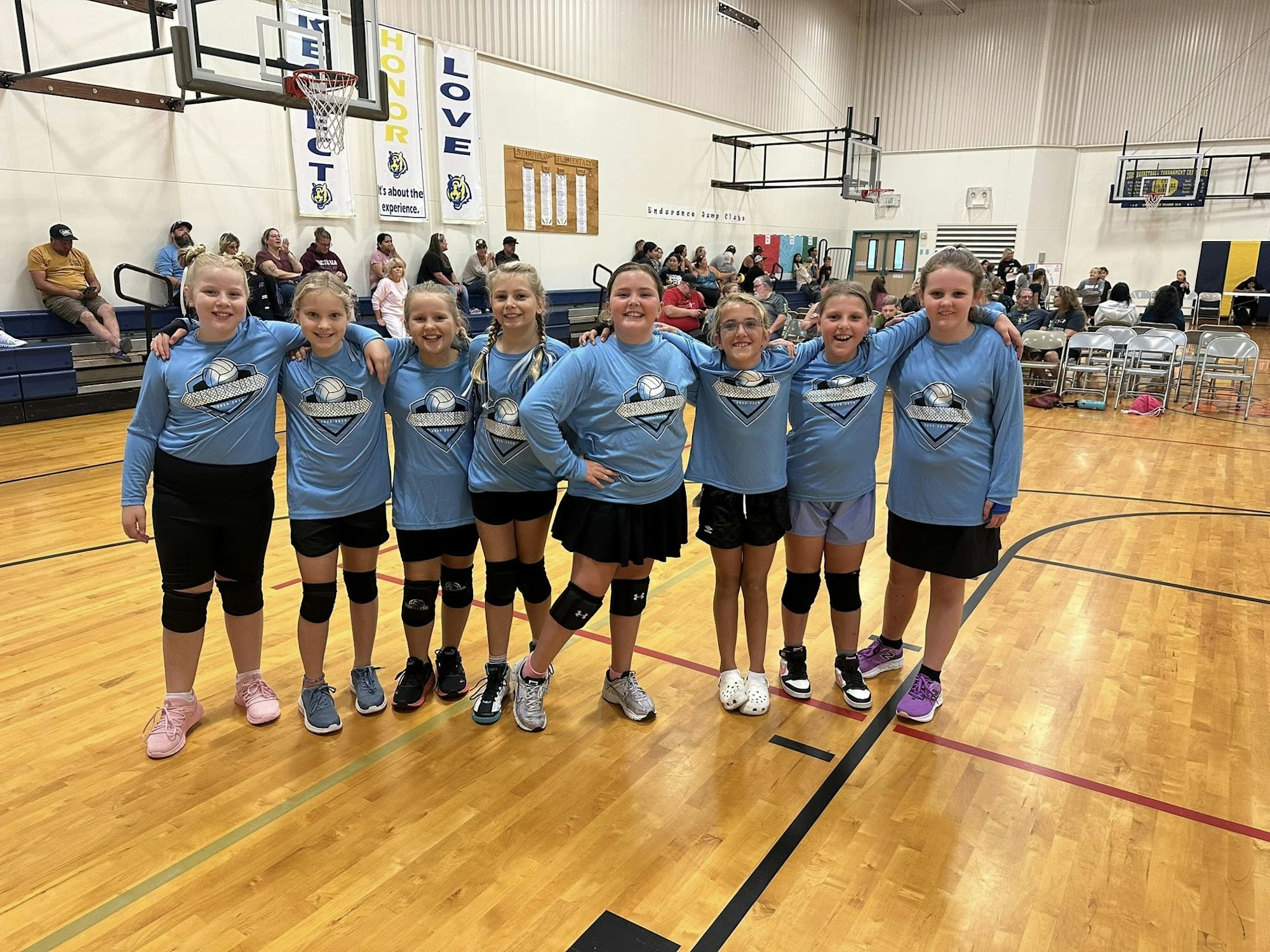 A group of young girls in matching blue volleyball shirts poses together on a gym court, smiling happily.