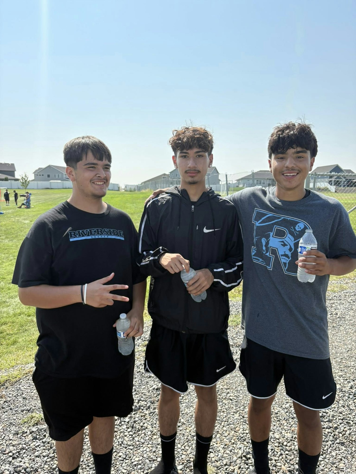 Three young men are standing together outdoors, holding water bottles and smiling. They are dressed in athletic wear.