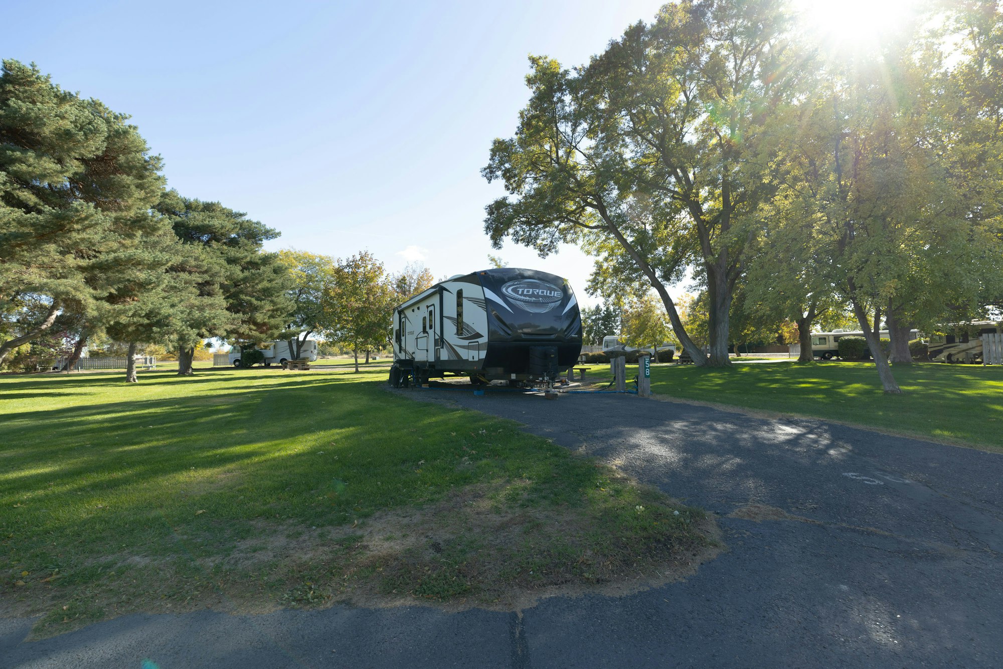A campsite with a large RV parked on green grass surrounded by trees on a sunny day.