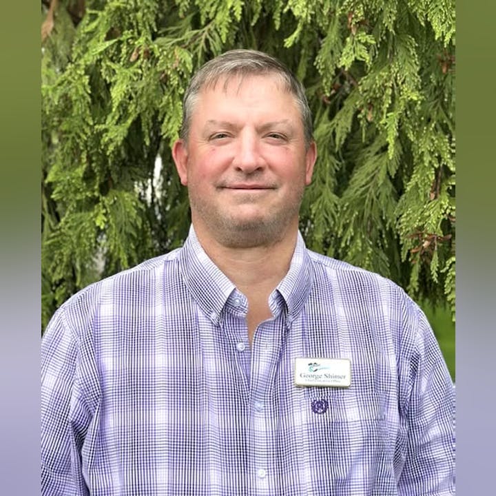 A man with short hair wearing a checkered shirt stands in front of lush greenery, looking at the camera.