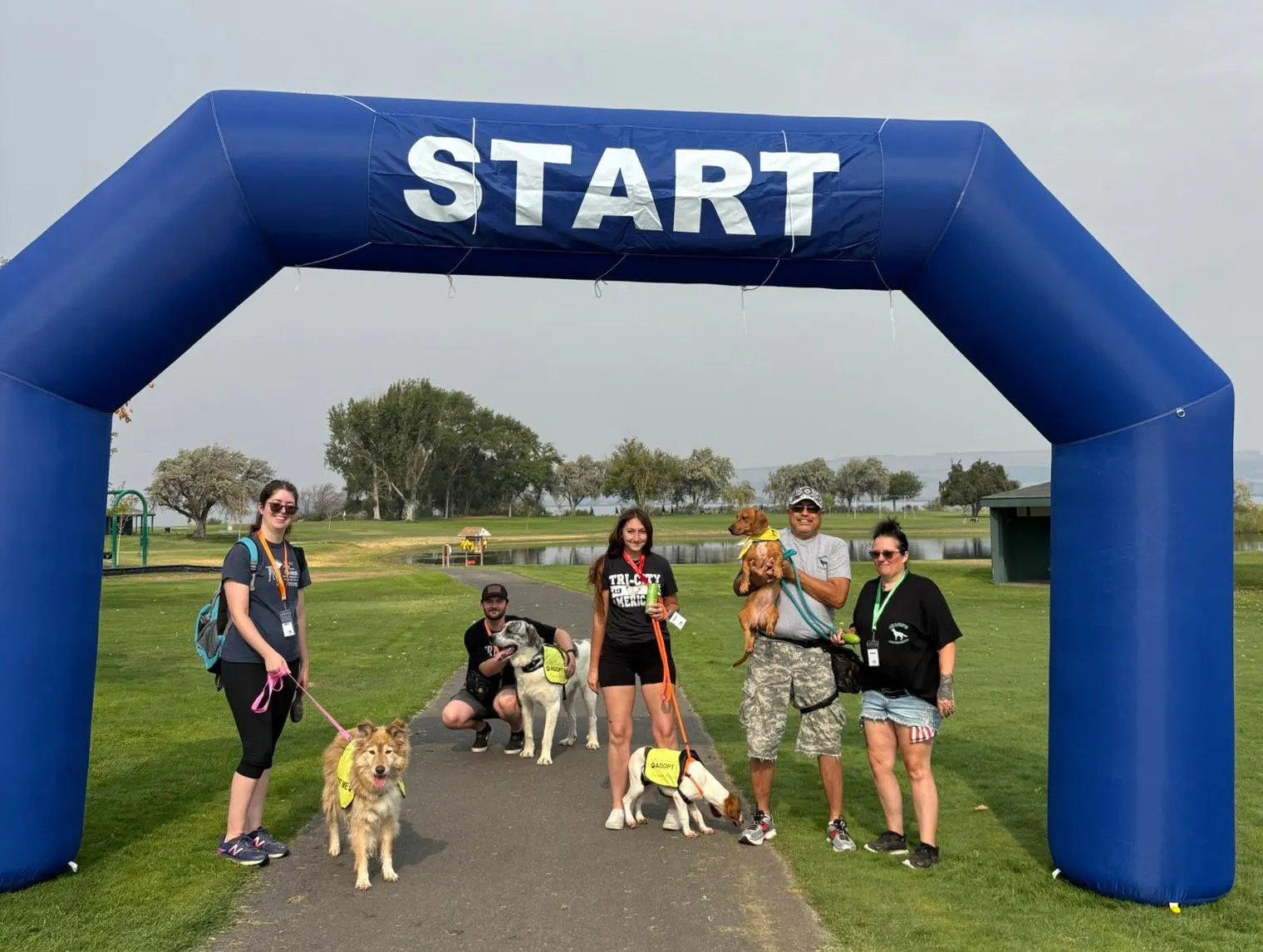 A group of people and dogs stand under a blue "START" arch at a park, likely preparing for a race or event.