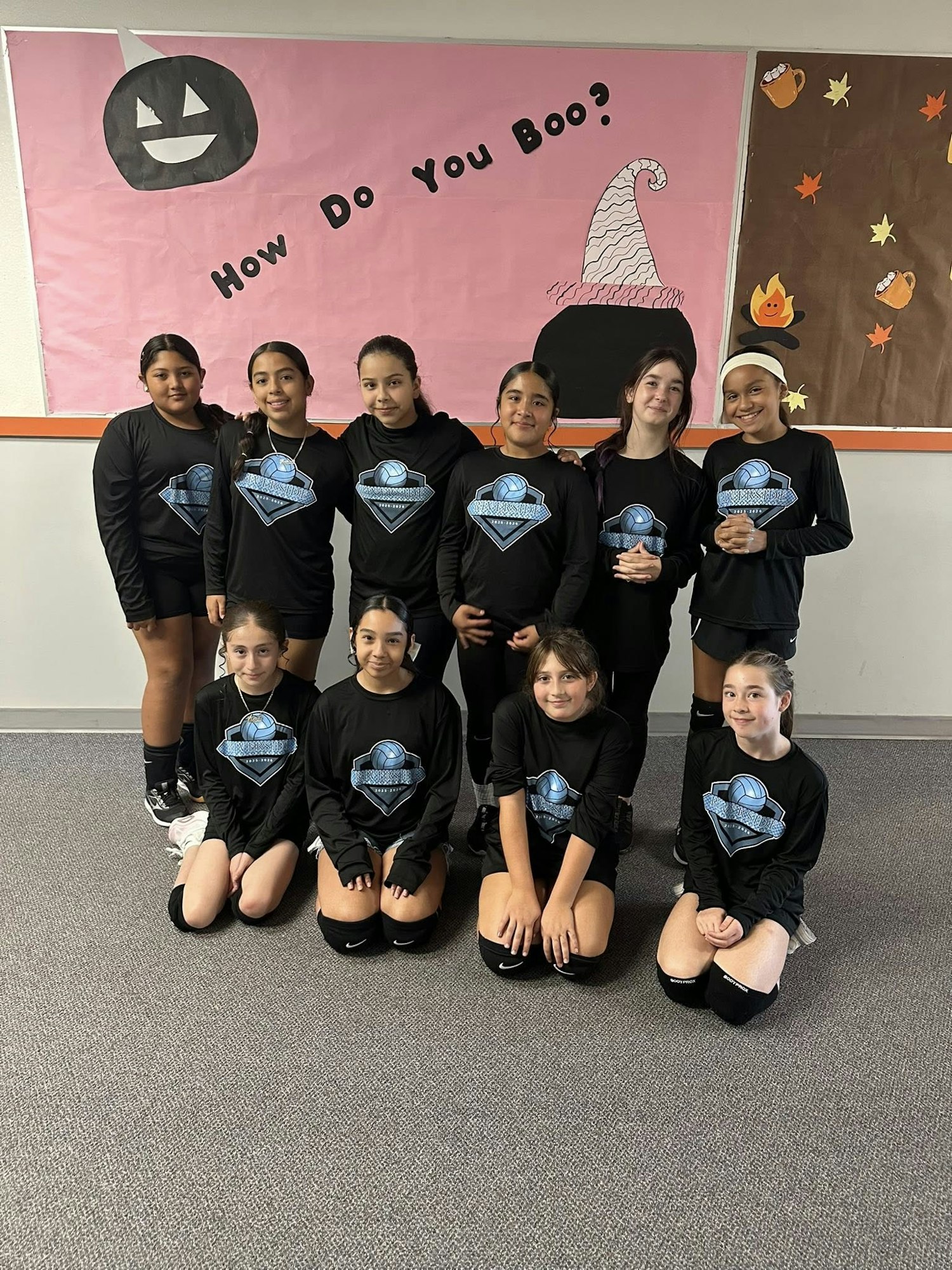 A group of young girls in matching black athletic shirts poses for a photo in front of a festive Halloween-themed backdrop.