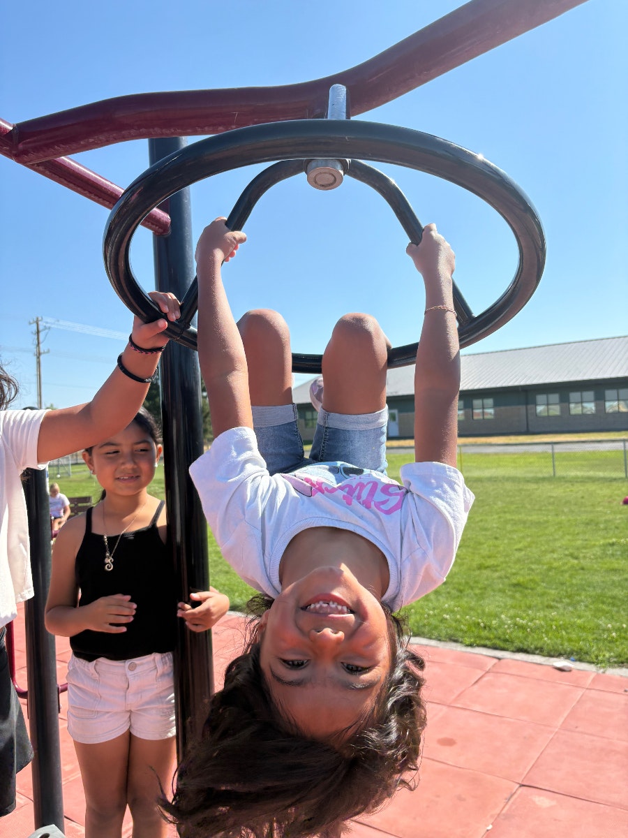 A sunny playground scene with kids; one girl joyfully hangs upside down from a play structure, smiling at the camera.