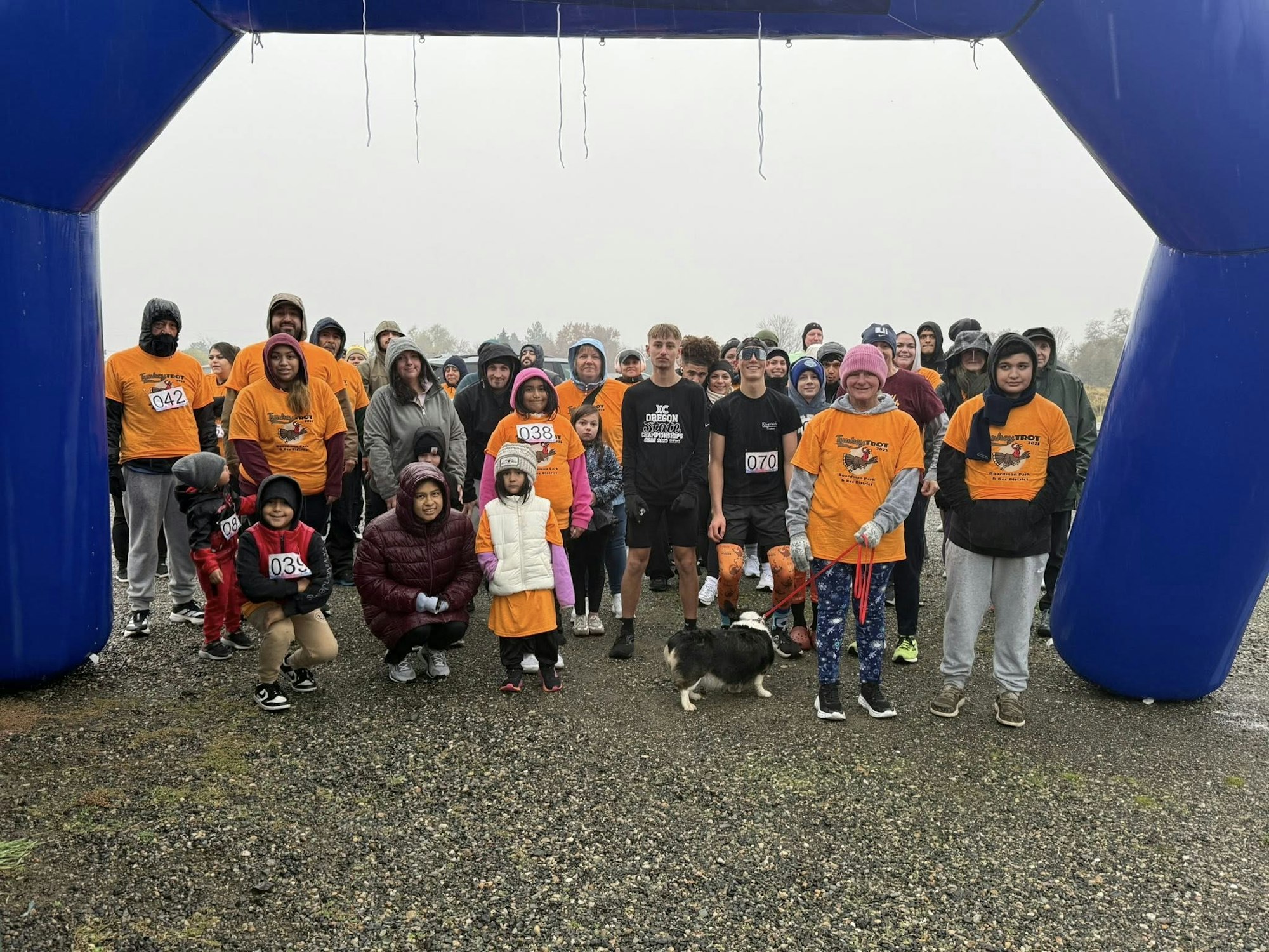 A group of people in orange shirts poses under a blue arch, likely preparing for a rainy community run or event.